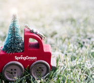 red toy truck with a Christmas tree in the trunk lit up with lights driving on a snowy lawn