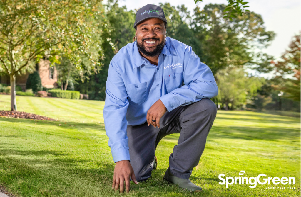 Pest control tech kneeling on the grass outside touching the ground, smiling big in SpringGreen uniform.