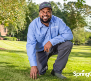 Pest control tech kneeling on the grass outside touching the ground, smiling big in SpringGreen uniform.